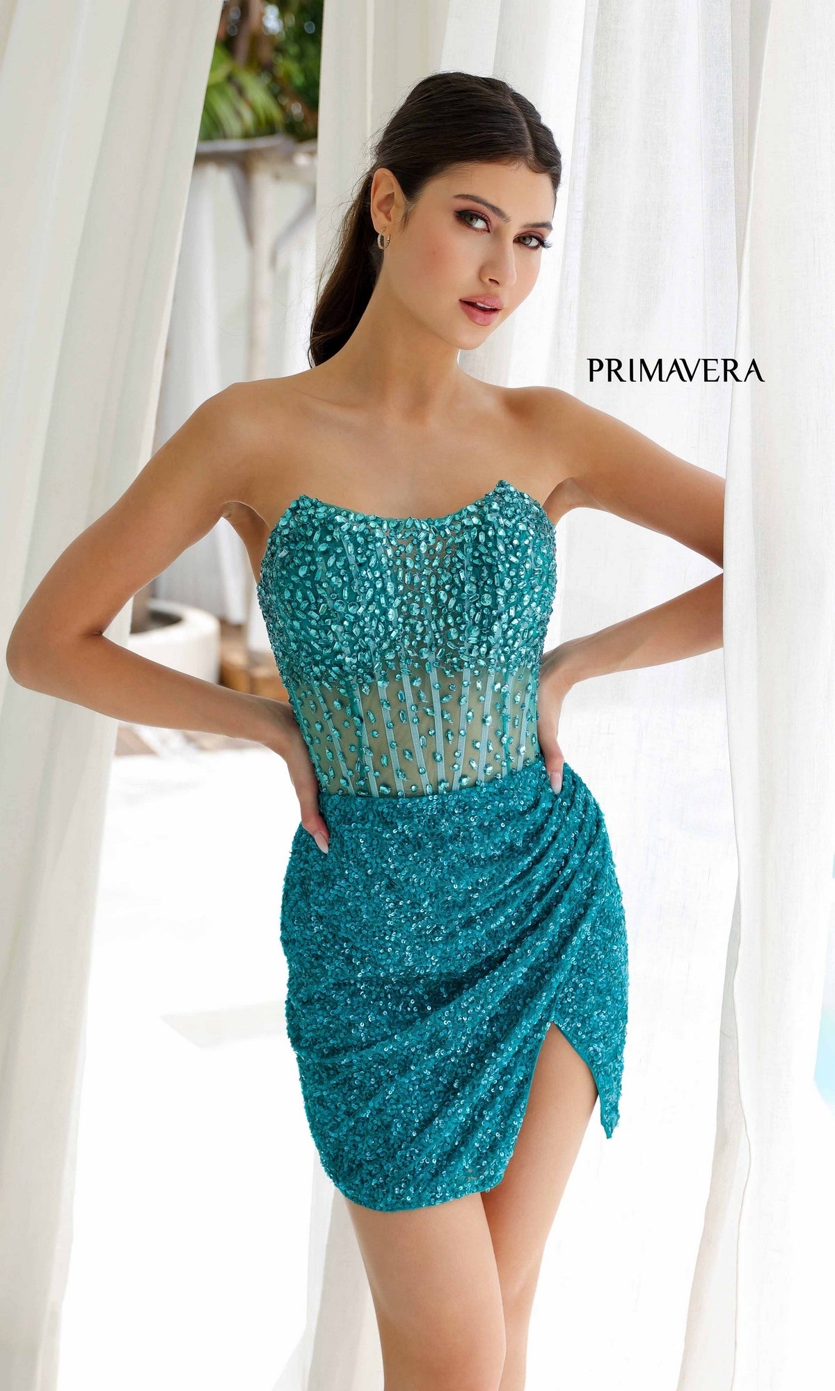 A woman in a teal, strapless sequin dress poses by white curtains with